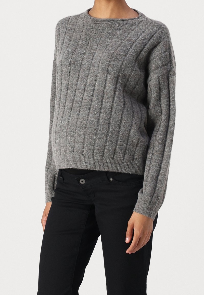 Woman wearing a grey knitted ribbed sweater with long sleeves and black pants, standing against a white background.