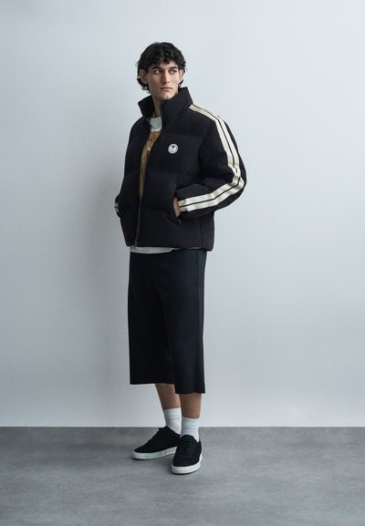 Νεαρός άνδρας με τα χέρια στις τσέπες, φοράει μαύρο μπουφάν με άσπρες ρίγες, μαύρο φαρδύ σορτς, άσπρες κάλτσες και μαύρα αθλητικά παπούτσια, στέκεται.