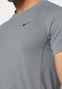 Harmaa lyhythihainen urheilupaita, jossa on kudottu verkkokuvio sivussa ja musta Nike-logo rinnassa.