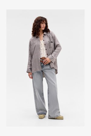 Jonge vrouw met krullend haar, gekleed in een oversized geruit overhemd, een witte kanten crop top, losse lichtblauwe jeans, beige schoenen en ringen om haar vingers.