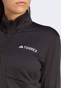 Kobieta w czarnej kurtce na zamek z wysokim kołnierzem, z białym logo "TERREX" po lewej stronie klatki piersiowej.