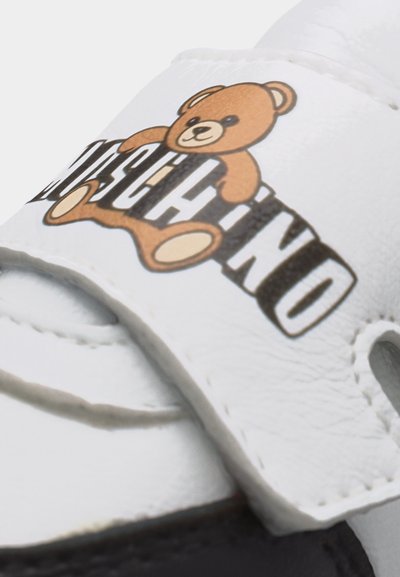 Λευκό λουράκι παπουτσιού με γραφικό καφέ αρκουδάκι πάνω από έντονο μαύρο και λευκό κείμενο "MOSCHINO" σε λείο δερμάτινο υπόβαθρο.
