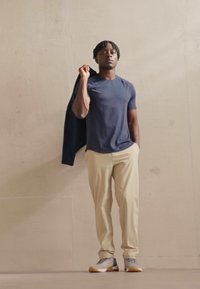 Marineblaues T-Shirt, beige Hose, graue Sneaker mit roten Akzenten und eine dunkle Jacke über einer Schulter. Glattes Material, schlanker Schnitt.