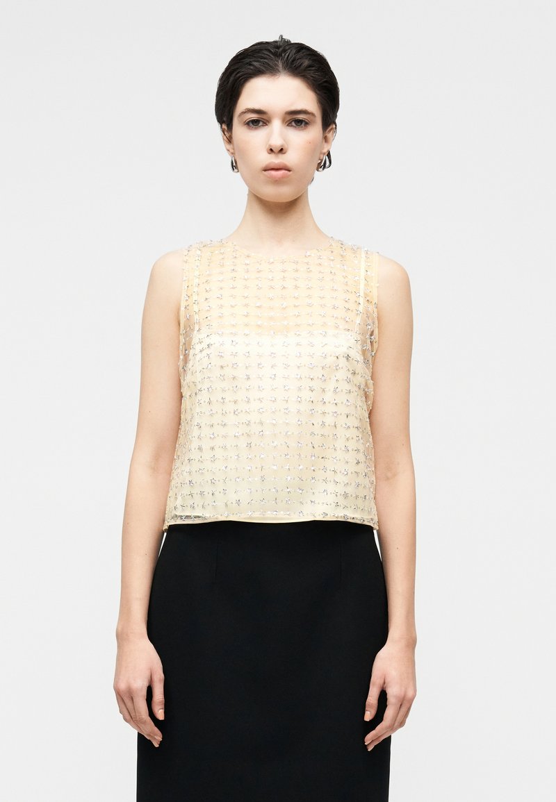 Bezrękawnik typu crop top wykonany z przezroczystej żółtej tkaniny, ozdobiony wzorami z srebrnych koralików, z okrągłym dekoltem i prostym dołem.