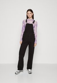 Black denim overalls with adjustable straps, large front pocket, silver hardware, and wide leg design, paired with a sheer lavender top.