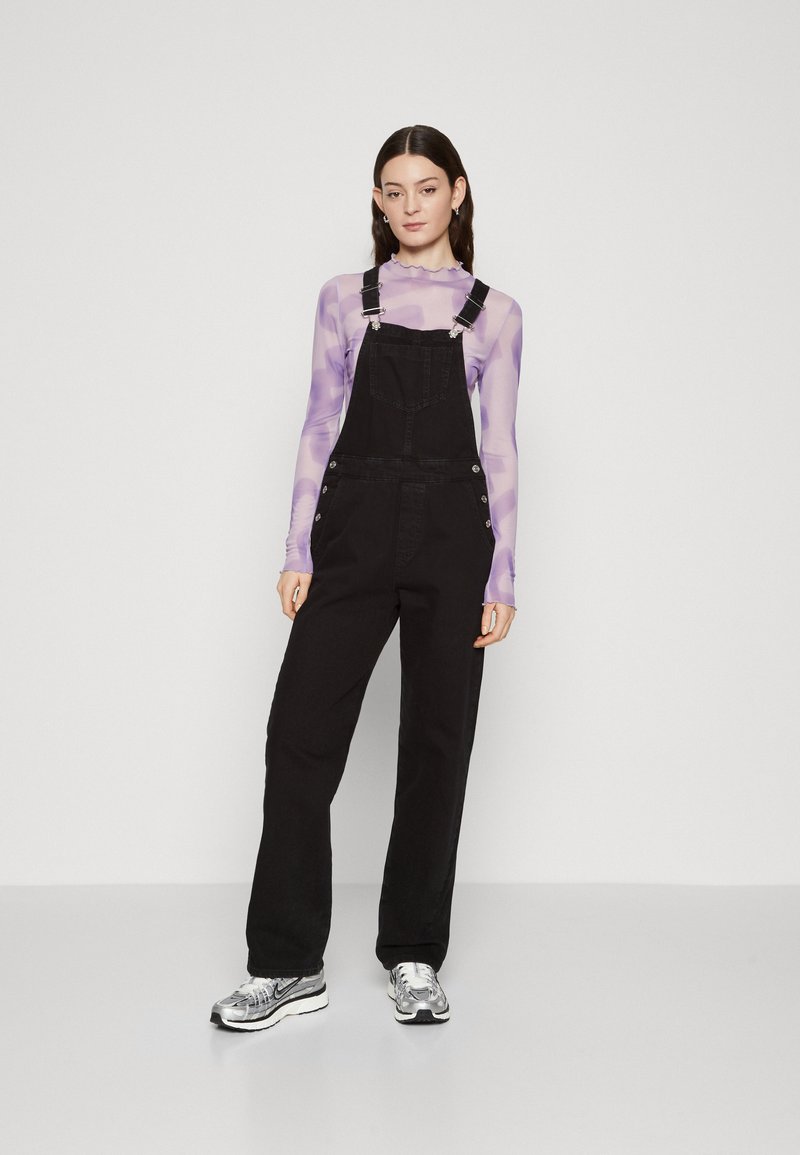 Black denim overalls with adjustable straps, large front pocket, silver hardware, and wide leg design, paired with a sheer lavender top.