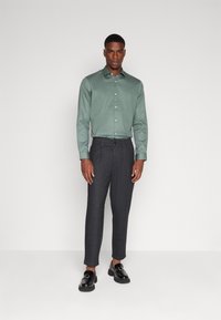 Chemise verte à boutons avec des manches longues et une texture lisse, associée à un pantalon à motifs sombres et des chaussures noires élégantes.