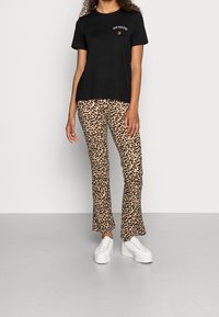 Vrouw in een zwart "GO NUTS!" t-shirt, beige en zwart luipaardprint broek en witte plateau sneakers die op een grijze vloer staat.