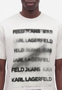 Άτομο που φοράει λευκό T-shirt με επαναλαμβανόμενο θολό και καθαρό μαύρο κείμενο ""KARL LAGERFELD JEANS"" μπροστά.