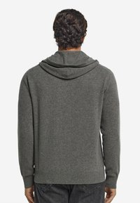 Man med mörkt lockigt hår som bär en grå hoodie och mörka jeans, vänd bort från kameran mot en enkel vit bakgrund.