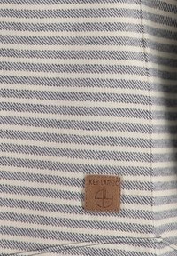 Tissu rayé en blanc et bleu marine, avec une surface texturée. Petite étiquette en cuir marron avec le logo de la marque cousue sur le tissu.