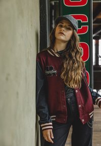 Bordeaux varsity jack met zwarte mouwen, met geborduurd "Revolution" logo. Draagt een grijze pet en zwarte broek. Lang golvend haar.