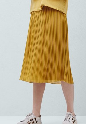 Femme portant une jupe midi plissée jaune moutarde et des baskets beige clair avec des accents imprimés léopard, debout devant un fond uni.