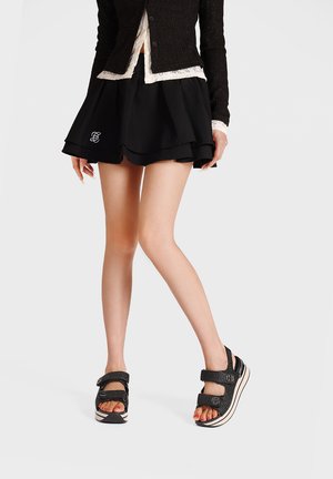 Donna con minigonna nera a strati con logo ricamato, sandali neri con texture, punta aperta e cinturini, e cardigan nero con bordatura in pizzo bianco.