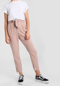 Vaaleanpunaiset räätälöidyt housut, joissa on sidottava vyötärönauha; yhdistetty valkoiseen lyhythihaiseen t-paitaan ja mustiin lenkkareihin. Sileä kangas, kapea istuvuus.