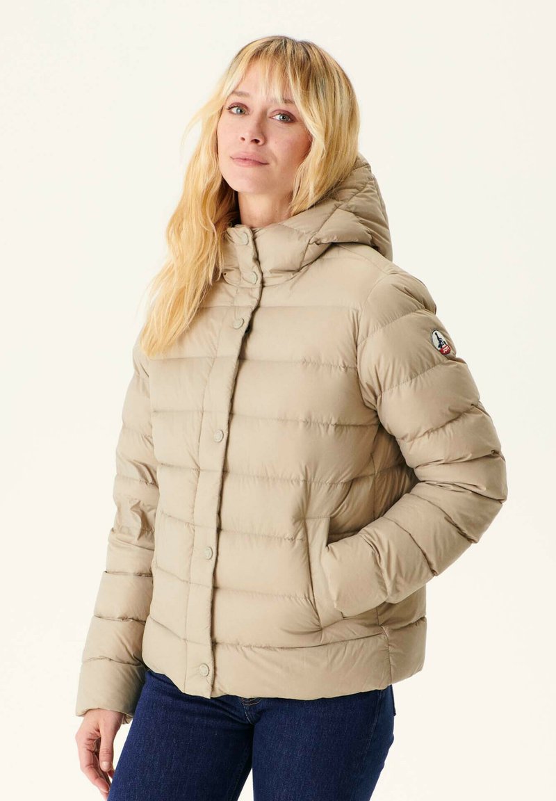 Chaqueta acolchada beige con capucha, que presenta un acolchado horizontal, botones a presión y un parche de logo en la manga. Textura suave y diseño informal.