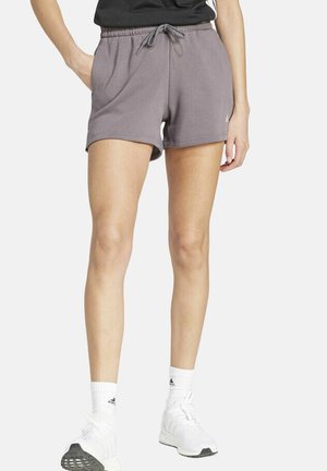 Frau trägt graue Shorts mit Kordelzug, weiße Sportsocken und weiße Turnschuhe, steht mit einer Hand in der Tasche vor weißem Hintergrund.