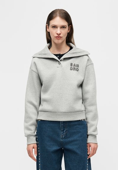 Egyenes barna hajú nő, szürke pulóvert visel, melyen a "SANDRO" felirat olvasható a mellkasán, valamint kék farmert oldalán szegecses díszítéssel, egy egyszerű háttér előtt áll.