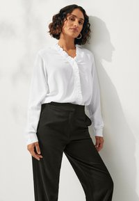 Witte blouse met lange mouwen en een ruffled V-hals, gecombineerd met hooggepolsterde zwarte broek. Stof is glad en de pasvorm is klassiek met knoopaccenten.