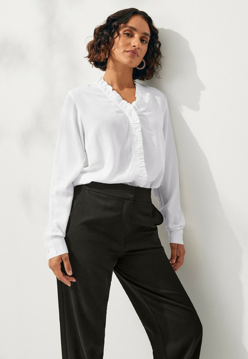Witte blouse met lange mouwen en een ruffled V-hals, gecombineerd met hooggepolsterde zwarte broek. Stof is glad en de pasvorm is klassiek met knoopaccenten.