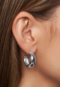 Pendiente de aro de metal plateado con una superficie lisa y pulida, que presenta un diseño curvado y un acabado reflectante. Sostenido en el lóbulo de la oreja.