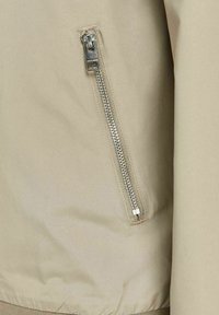 Béžová látka bundy s viditelným stříbrným zipem na levé straně. Hladká textura s přiléhajícím spodním lemem a bez dalších vzorů.