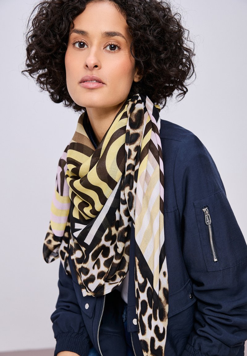Donna con capelli ricci scuri che indossa una sciarpa a motivi con stampa animalier e forme geometriche sopra una giacca blu navy con tasca sulla manica con zip.