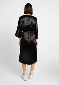 Musta satiinikangasmekko, jossa on kolmennes hihansuut, vyötärö on kiristetty ja helma on valuva. Mekossa on sileä tekstuuri ja polveen ulottuva helma.