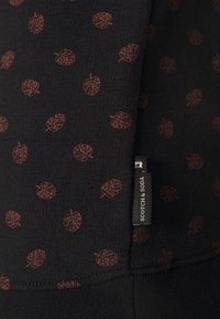 Black fabric featuring a repeating leaf pattern in burnt orange. Includes a small "Scotch & Soda" tag at the edge. Soft, textured material.