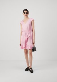 Pink dress with a V-neck, short cap sleeves, and a ruffled hem. Features an elasticized waist and a small black handbag. Flat black shoes.