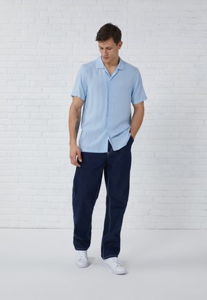 Man in lichtblauw shirt met korte mouwen, donkerblauwe broek en witte sneakers staand tegen een witte bakstenen muur, kijkt naar beneden, één hand in de zak.