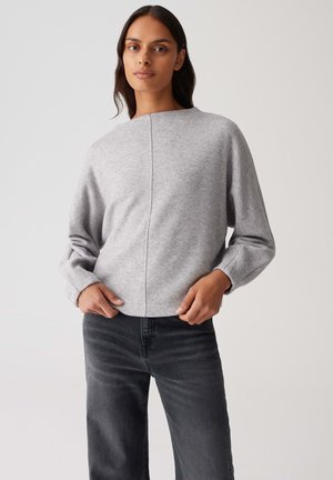 Femme portant un pull à manches longues gris clair et un jean noir taille haute, se tenant avec les mains près des poches devant un fond uni.
