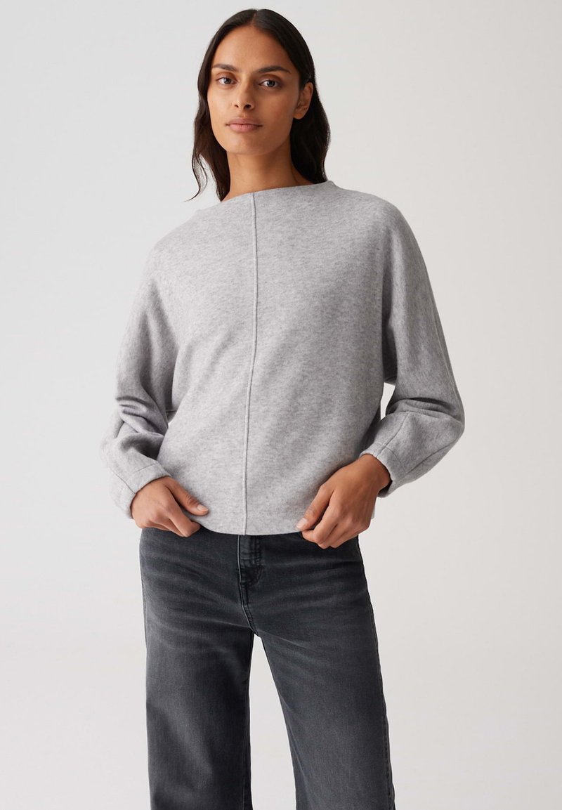 Donna che indossa un maglione grigio chiaro a maniche lunghe e jeans scuri a vita alta, in piedi con le mani vicino alle tasche su uno sfondo semplice.