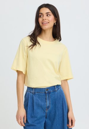 Ung kvinde smilende, iført en lysegul kortærmet skjorte stoppet ned i højhælede blå denim bukser med plisseringer, stående mod en ensfarvet baggrund.