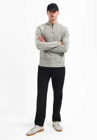 Lichtgrijs gebreid sweater met een kwart rits, gecombineerd met zwarte broek en grijze sneakers met witte strepen. Een zwarte pet maakt de look compleet.