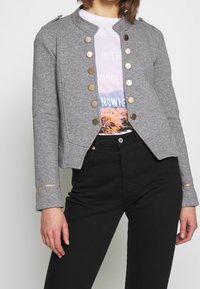 Giacca grigia corta con polsini e bottoni dorati. Abbinata a una t-shirt grafica, pantaloni neri aderenti completano il look.