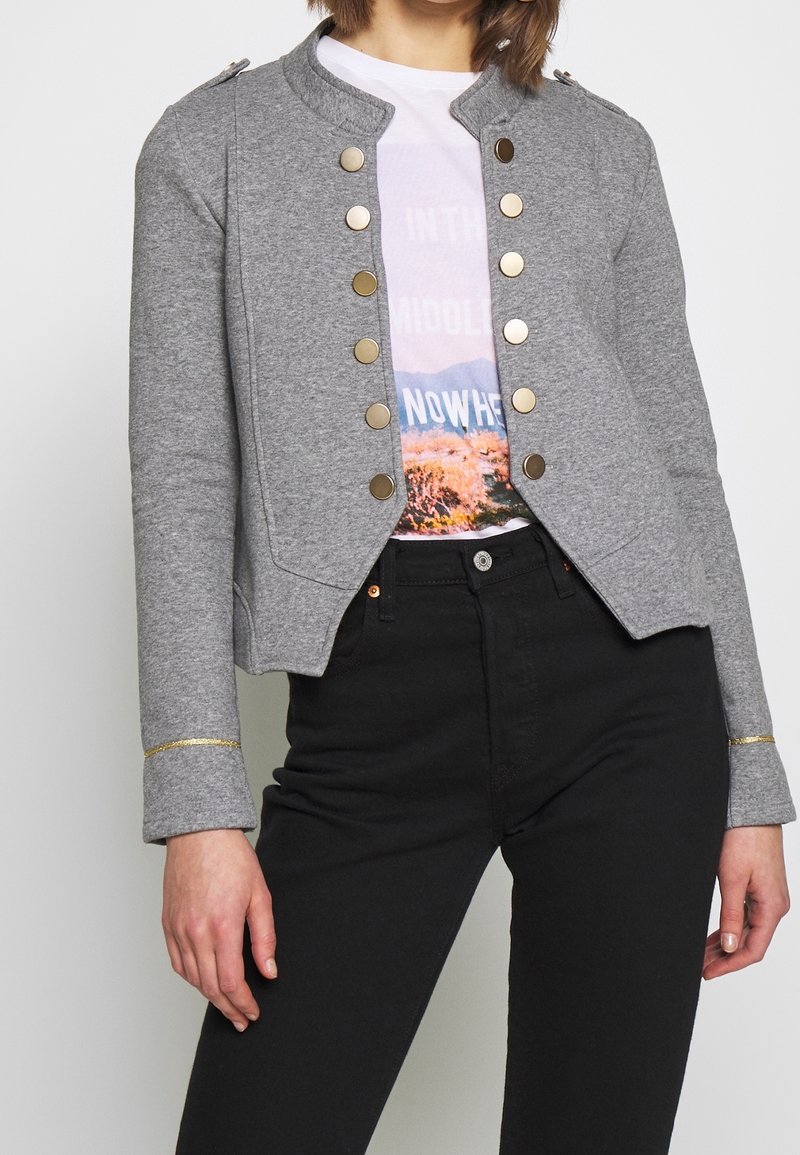 Giacca grigia corta con polsini e bottoni dorati. Abbinata a una t-shirt grafica, pantaloni neri aderenti completano il look.