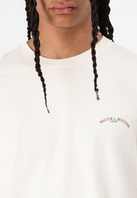 Light beige cotton t-shirt with a round neckline. Features a small embroidered logo in dark red and green on the left chest area.