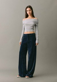 Nainen seisoo päällään harmaa olkaimeton lyhyt villapaita, leveälahkeiset tummansinsiset samettihousut ja valkoiset avonaiset sandaalit yksinkertaista taustaa vasten.