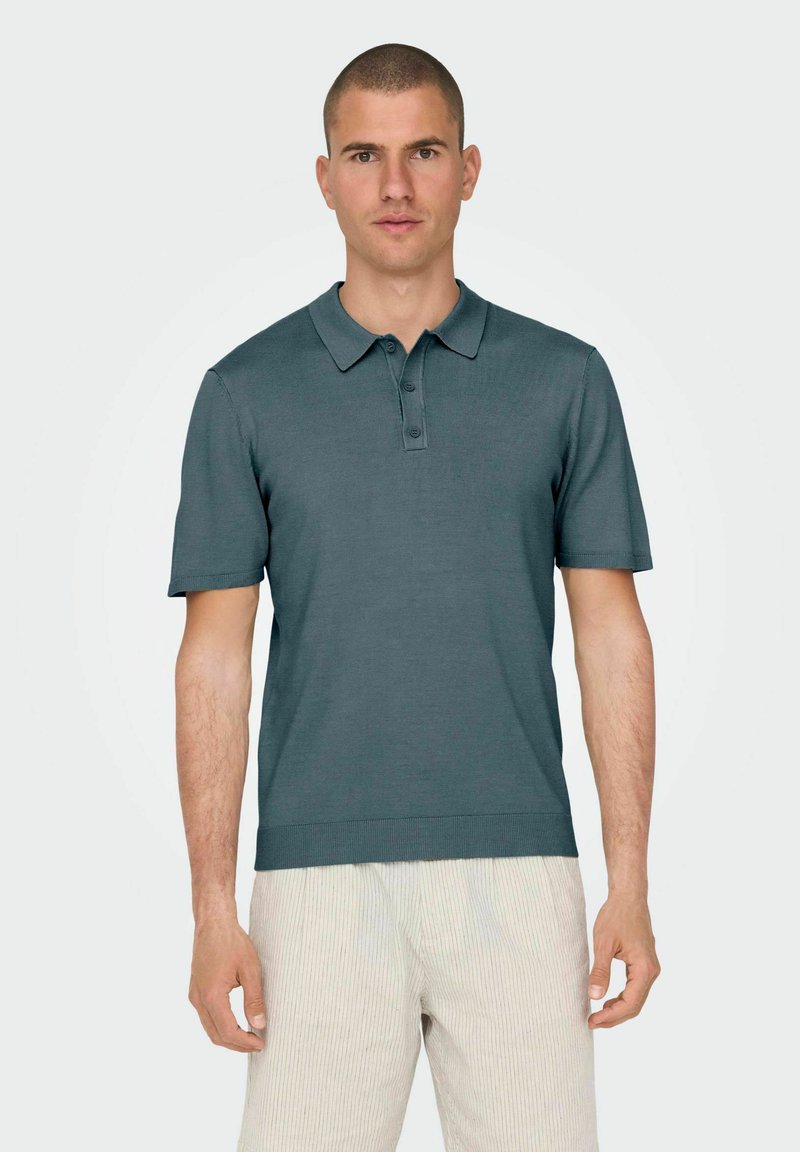 Polo a maniche corte di colore teal scuro, realizzato in tessuto liscio. Presenta un colletto classico, una pattina con bottoni e orli a costine.