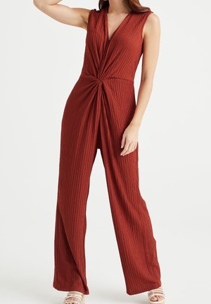 Mujer que lleva un mono sin mangas de color óxido texturizado con un detalle de torcido en la parte delantera, combinado con sandalias blancas de tiras, de pie frente a un fondo blanco.