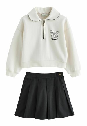 Jong meisje met lang krullend haar draagt een wit sweatvest met rits en een zwarte geplooide rok, staand op een lichtgrijze vloer en witte achtergrond.