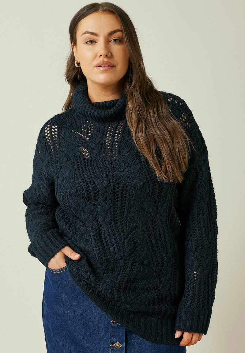 Dunkelblau gestrickter Pullover mit hohem Kragen, der ein strukturiertes Zopfmuster und gerippte Bündchen aufweist und über einem Jeanskleidungsstück getragen wird.
