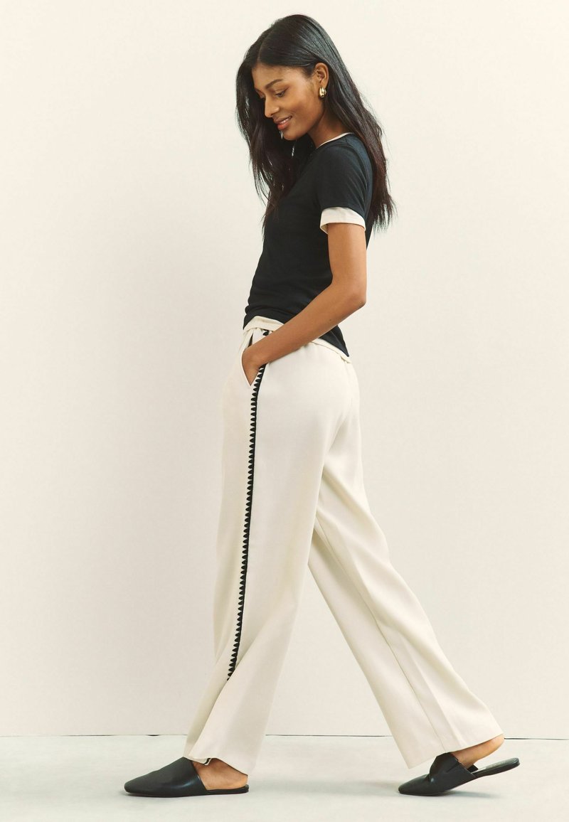 Femme marchant portant un haut noir à manches courtes, un pantalon crème à jambes larges avec une bordure noire sur le côté, et des chaussures noires sans lacets, souriante les mains dans les poches.