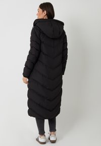 Zwarte pufferjas met een chevronpatroon, lange lengte, capuchon en elastische manchetten. Draagt met donkere jeans en lichtgekleurde sneakers.