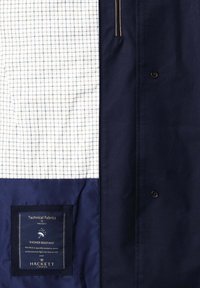 Vnútro námorníckeho saka so vzorovanou bielou kockovanou podšívkou, vreckom na zips, čiernymi gombíkmi a štítkom s pokynmi na starostlivosť o tkaninu, ktorý uvádza, že ide o sprchovo odolnú technickú látku.