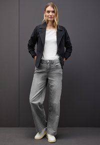 Chaqueta negra estructurada sobre una camisa blanca, combinada con jeans grises sueltos y deslavados, y zapatillas blancas. El fondo es de un color oscuro sólido.