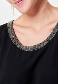 Top nero con scollatura arrotondata decorato con piccole perline metalliche, creando un accento testurizzato intorno al collo. Tessuto liscio e leggero.