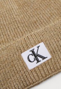 Tejido de lana de punto beige con una etiqueta rectangular blanca que presenta el logotipo "cK" en negro y en letras negritas.