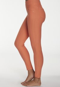 Bare feet wearing fitted, high-waisted, rust-colored leggings standing sideways against a plain background.